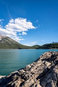 Rocky shore along a calm blue lake with forested hills and mountains under a blue sky.