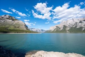 Calm blue lake surrounded by green and rocky mountains under a blue sky with white clouds.