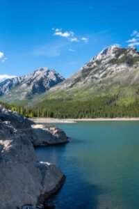 Mountain lake with rocky shoreline, evergreen forest, and clear blue sky with clouds.