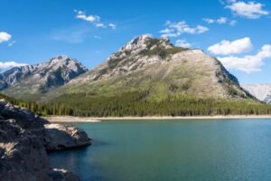 Lake with rocky shore and forest, mountainous terrain under blue sky with clouds.
