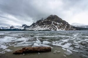Partially frozen lake with floating ice and a log, surrounded by snow-covered mountains under cloudy sky.