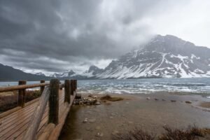 Wooden pier over partially frozen lake with snow-covered mountains under cloudy sky.