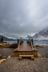 Wooden pier over icy water with snowy mountains and cloudy sky in the background.