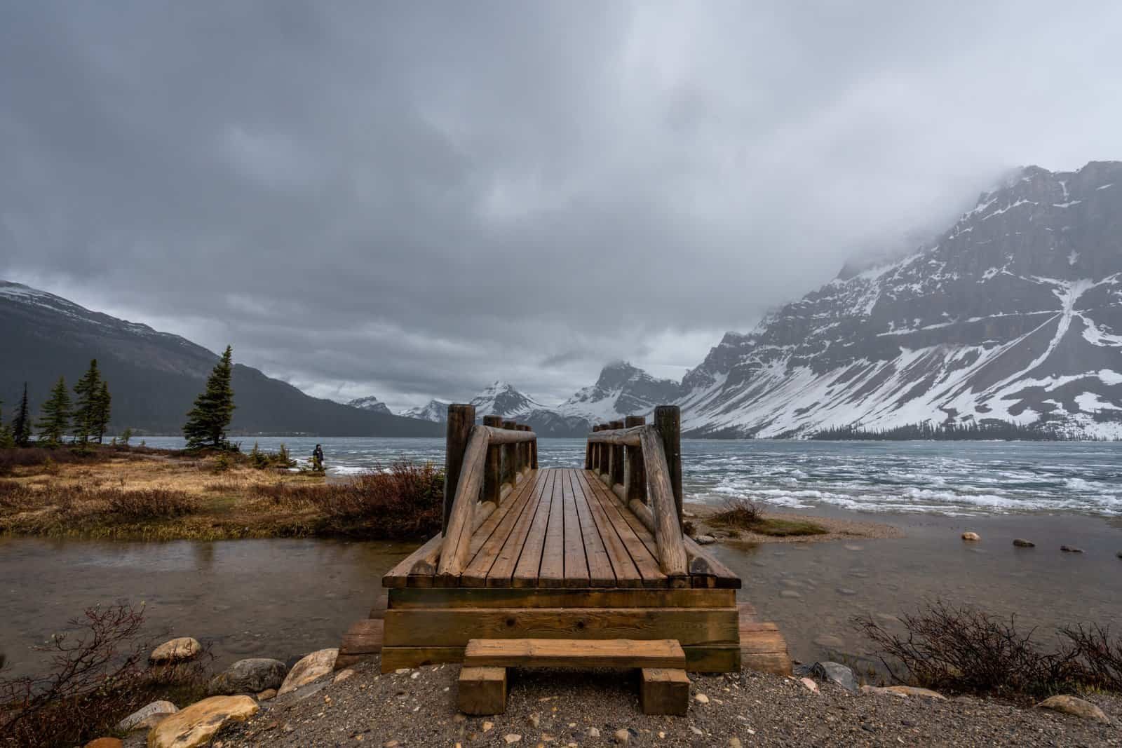 Wooden bridge over small water body leading to partially frozen lake with snow-covered mountains.