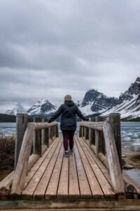 Person wearing winter clothing standing on wooden bridge overlooking icy lake and snow-covered mountains under cloudy sky