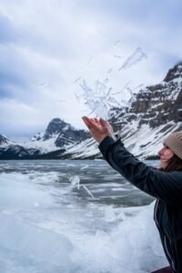 Person in winter clothing tossing ice shards near frozen lake with snow-covered mountains.