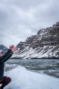 Person in dark jacket throws snow by frozen lake with snow-covered mountains under cloudy sky