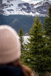 Person wearing beige knit hat looks at lake, evergreen trees, snow-covered mountain, and red building.