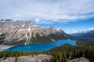 Bright blue lake surrounded by evergreen forest and snow-capped mountains under a cloudy sky