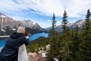 Person in black jacket and beige hat photographing blue lake, evergreen trees, and snow-capped mountains.