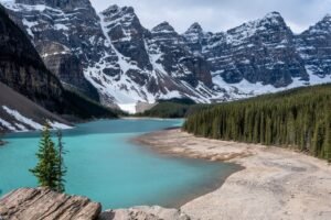 Turquoise glacial lake beside rocky shore and evergreen forest with snow-capped mountains.