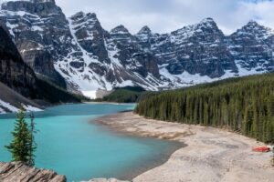 Turquoise lake with rocky snow-capped mountains, dense forest, and sandy shore with boats.