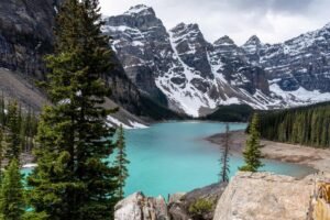 Turquoise lake surrounded by evergreen trees, rocky terrain, and snow-capped mountains under cloudy sky