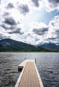 Wooden dock on a lake with forested mountains and snow-capped peaks under a partly cloudy sky