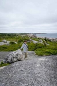 Person leaning on large rock on coastal rocky terrain with greenery and ocean in background
