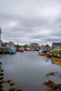 Small harbor with colorful fishing boats docked near wooden piers, buildings, and rocky shore under cloudy sky.