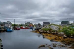 Coastal fishing village with boats docked, rocky seaweed shoreline, wooden houses, and overcast sky.