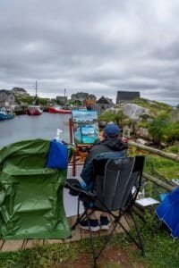 Person painting a harbor scene on an easel by a rocky waterfront with boats and houses.