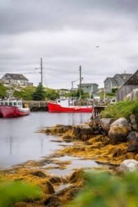 Harbor with two red fishing boats, rocky seaweed-covered shore, houses, and cloudy sky.