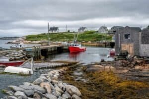 Harbor with a red fishing boat docked by pier, rocky shore, wooden buildings, and houses on hill.