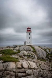 White lighthouse with red top on rocky shore under cloudy sky by the ocean.