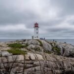 White lighthouse with red top on rocky coast overlooking ocean under cloudy sky with people nearby