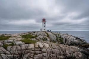 White lighthouse with red top on rocky coast overlooking ocean under cloudy sky with people nearby