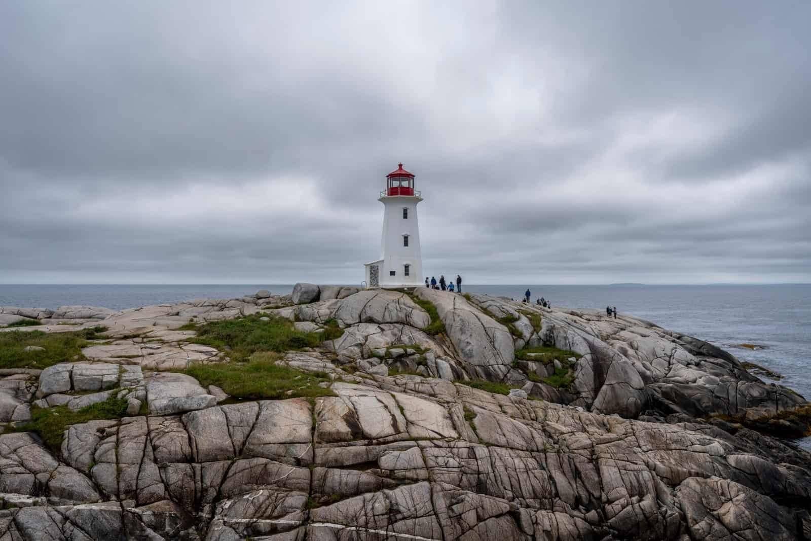 White lighthouse with red top on rocky coast overlooking ocean under cloudy sky with people nearby