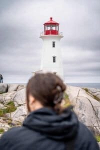 Person in dark jacket facing a white lighthouse with red top on rocky shore under cloudy sky