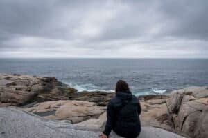 Person in dark jacket sitting on rocky shore facing calm ocean under cloudy sky.