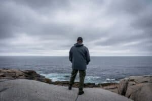 Person in dark jacket and green pants standing on rocky shore looking at ocean under cloudy sky