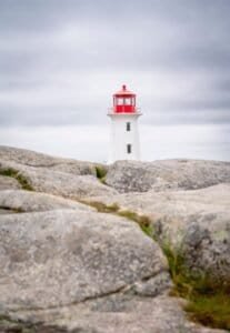 White lighthouse with red top behind large gray rocks under cloudy sky.