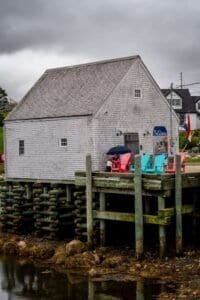 Gray shingled Neil Depew Gallery on wooden pier over rocky shore with colorful chairs and person under umbrella.
