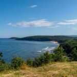 Coastal view with blue sea, sandy beach, green shrubs, forested hills, and a partly cloudy sky.