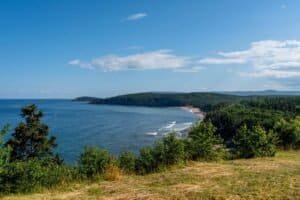 Coastal landscape with beach, forest, and rolling hills Coastal view with blue sea, sandy beach, green shrubs, forested hills, and a partly cloudy sky.