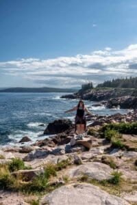 Woman standing on rock by rocky coastline with ocean and forest background Woman stands on rock by rocky coastline with ocean, waves, forest, and partly cloudy sky.