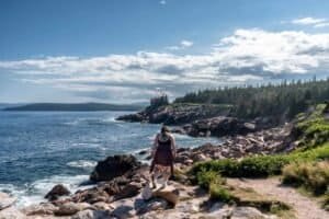 Person standing on rock by rocky coastline with pine forest background Person standing on rocky coastline with ocean waves and forested cliffs under a partly cloudy sky