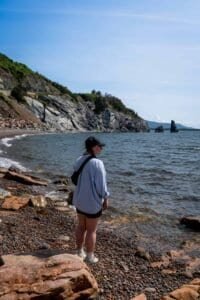 Person in light blue shirt and black cap stands on rocky shore facing the ocean with cliffs and rock formations.