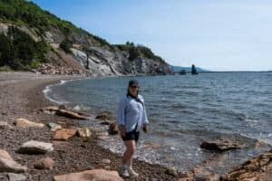 Person in blue shirt and black cap standing on rocky shoreline with cliffs and ocean rocks nearby.
