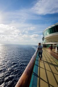 Person leaning on cruise ship railing overlooking ocean with others on deck and blue sky.