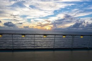 Ship deck railing with yellow lights overlooking the sea and sunset with partly cloudy sky.