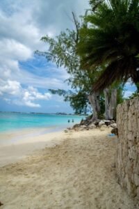 Sandy beach with turquoise water, trees, stone wall, people wading, and distant buildings under clouds.