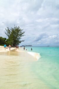 Beach with light sand, turquoise water, people walking and swimming, trees and cruise ships in distance.