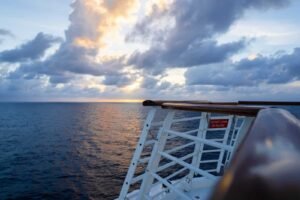 Ocean view at sunset from a ship deck with a white railing and a do not climb sign.