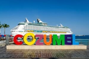 Large white cruise ship docked behind colorful Cozumel sign on wooden boardwalk.