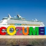 An image of a cruise ship with a sign that says Cozumel in the foreground with blue sky and a blue ocean