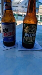Two beer bottles, Tecate Light and Indio, on napkins on a blue table near a docked cruise ship.