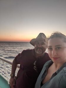Two people on a boat deck with ocean and sunset sky in the background.