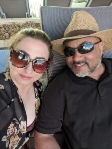 A man in a straw hat and black polo shirt and a woman in floral top and large sunglasses sitting outdoors.