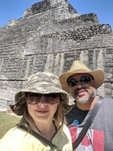 Two people wearing hats and sunglasses take a selfie in front of an ancient stone pyramid.